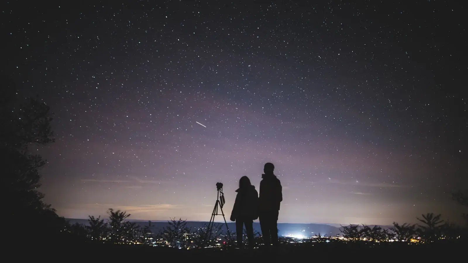 Two people beside a camera on a tripod watch a star-filled sky with a shooting star, above the lights of a valley