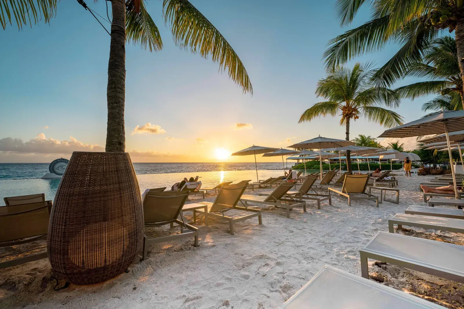Sunset over the Caribbean Sea seen from a palm-lined beach with loungers, parasols and an infinity pool