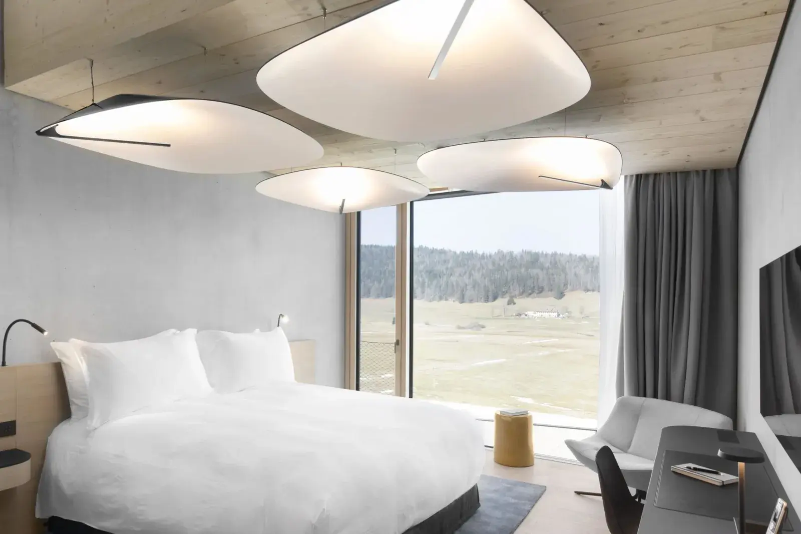 A hotel room with a timber ceiling and sculptural white pendant lamps, its floor-to-ceiling window opening onto a meadow and forest