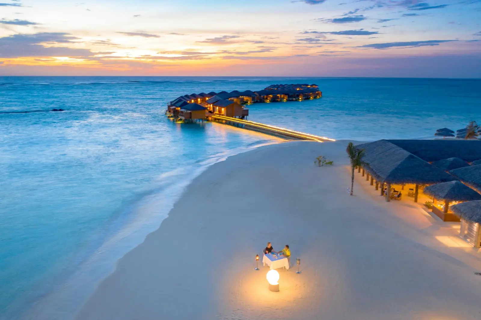 Aerial view at dusk of overwater villas linked by a lit jetty, a private candlelit dinner for two on a white-sand beach beside a turquoise lagoon