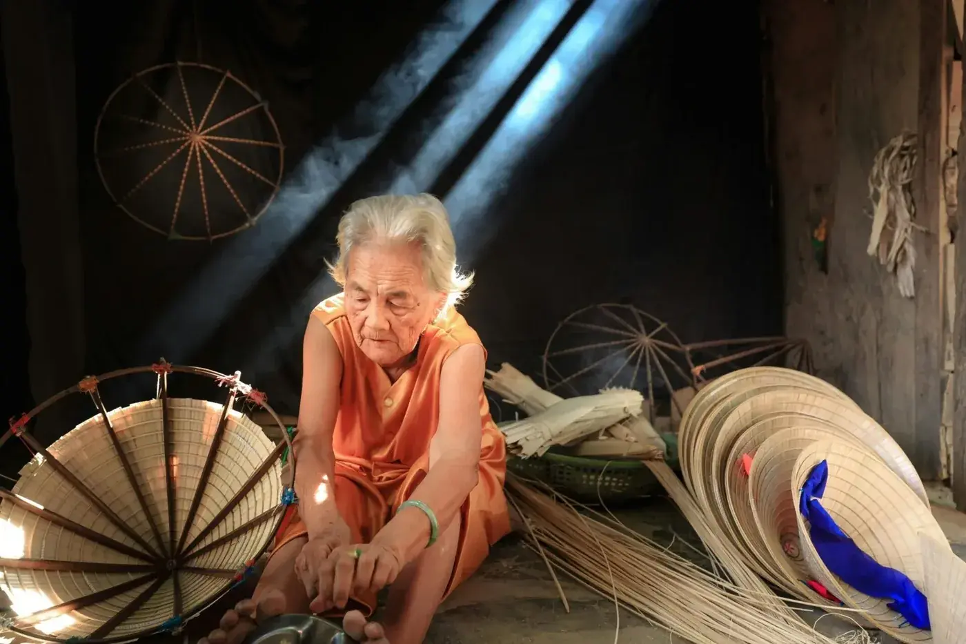 An elderly woman in an orange dress weaves conical palm-leaf hats in a dim workshop crossed by shafts of light