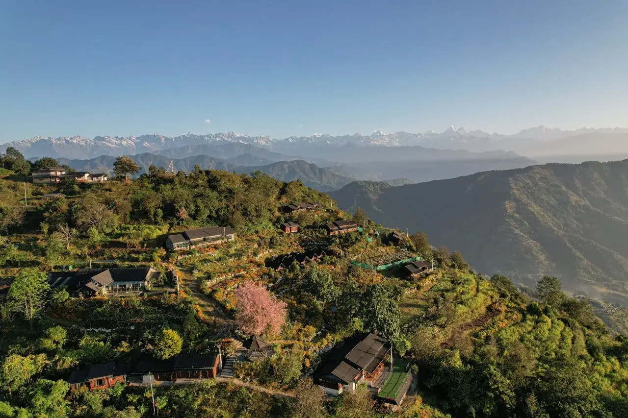 Aerial view of hillside lodges and terraced gardens on a green ridge, with the snow-capped Himalaya on the horizon