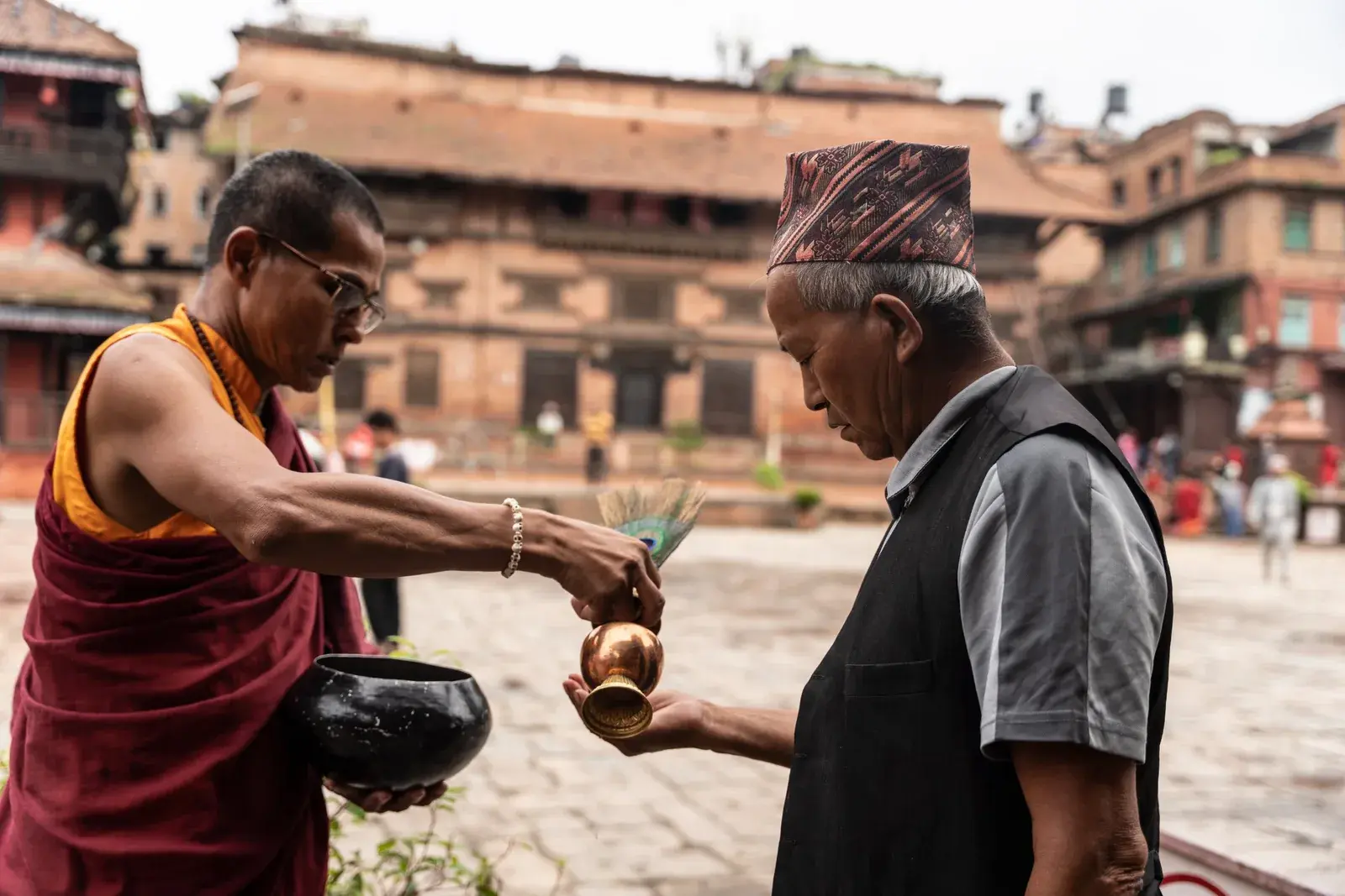 A monk in maroon and saffron robes pours from a copper vessel into the cupped hand of an older man in a traditional cap, in a brick-built heritage square