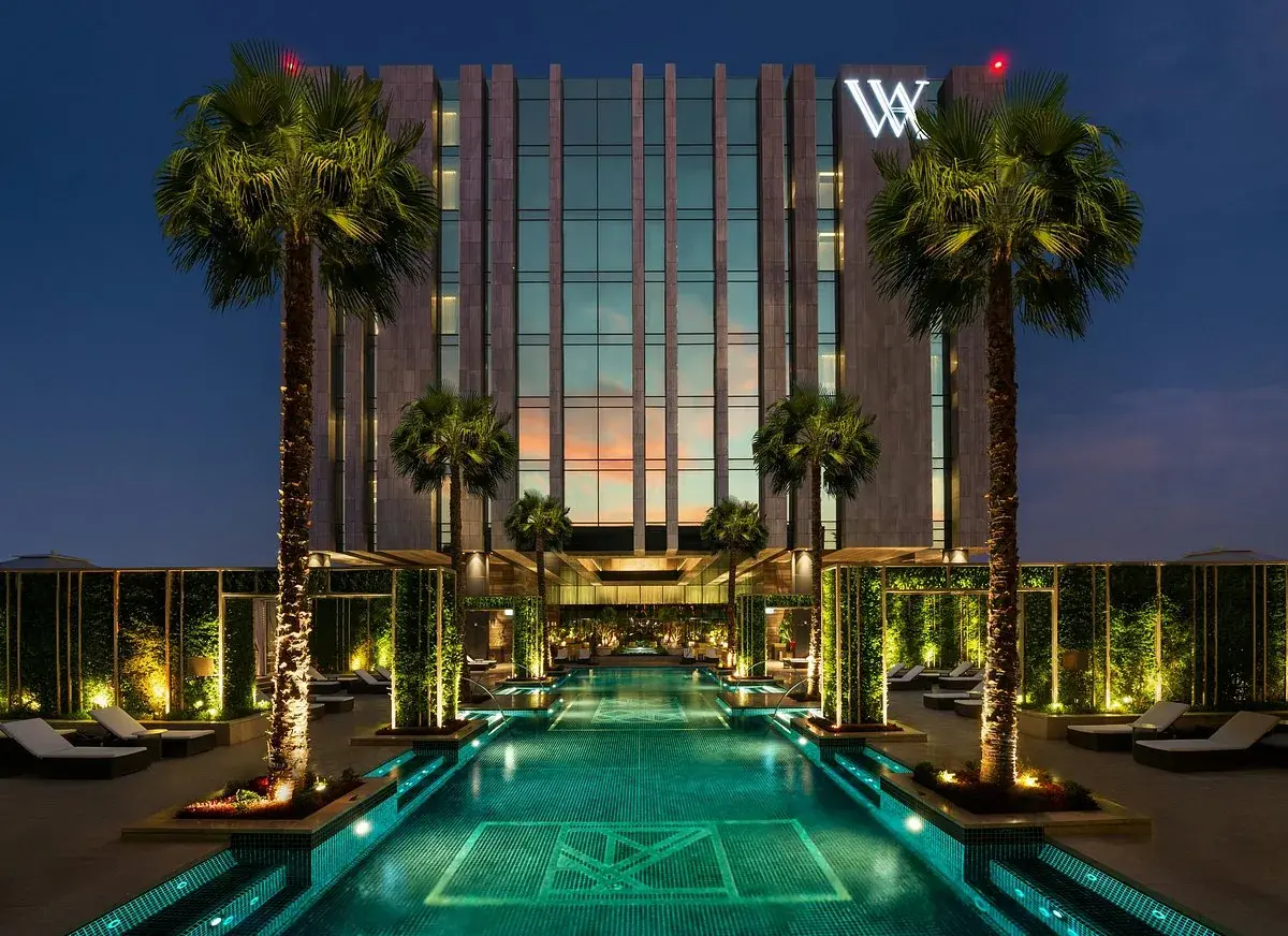 A long tiled pool lit at dusk, framed by palm trees and planted green walls, in front of a glass hotel tower
